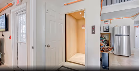 Interior view of a facility hallway showing an open elevator with wooden interior, a closed white door, a stainless steel refrigerator, and part of a decorated Christmas tree. The walls and ceiling have exposed orange pipes and there is an exit sign above a door with glass panels.