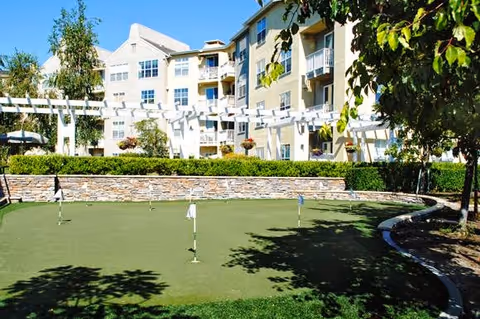 Outdoor putting green and landscaped courtyard in front of a multi-story senior living building with balconies and a pergola.