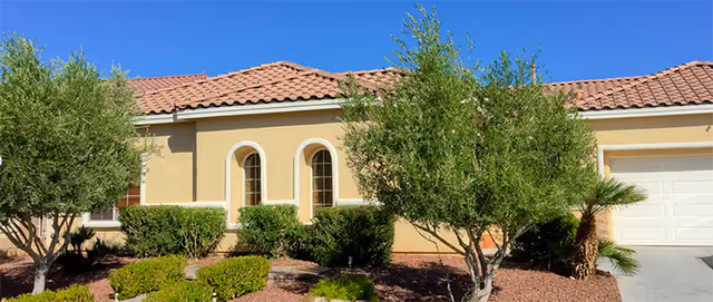 Front exterior view of a single-story house with a tan stucco finish and a red tile roof. The house has three arched windows and a white garage door. There are green shrubs and small trees in the landscaped front yard with brown mulch.