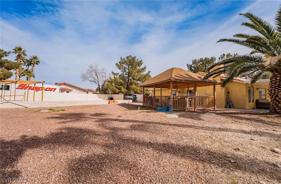 Outdoor area of a senior living facility featuring a gravel ground, a wooden gazebo with a roof, palm trees, and a yellow building in the background under a partly cloudy sky.