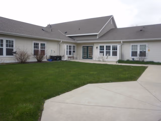 Exterior view of a single-story building with beige siding and multiple windows. There is a concrete pathway leading to a patio area with outdoor chairs and tables. The lawn is green and well-maintained under an overcast sky.