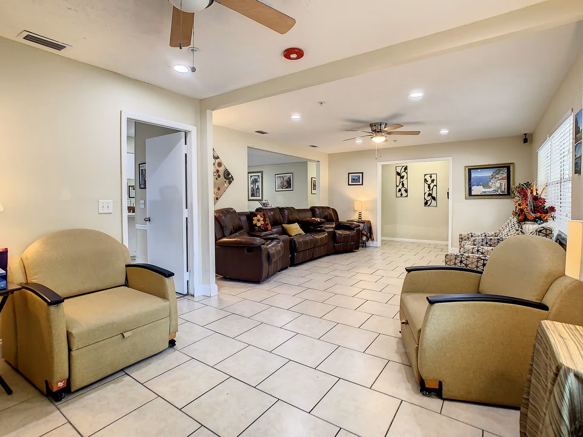 A spacious living room area with beige tiled floors and cream-colored walls. The room features two beige armchairs in the foreground, a brown leather reclining sofa against the far wall, and two patterned armchairs near a window with blinds. Ceiling fans and recessed lighting illuminate the space. Decorative artwork and a floral arrangement add color to the room.