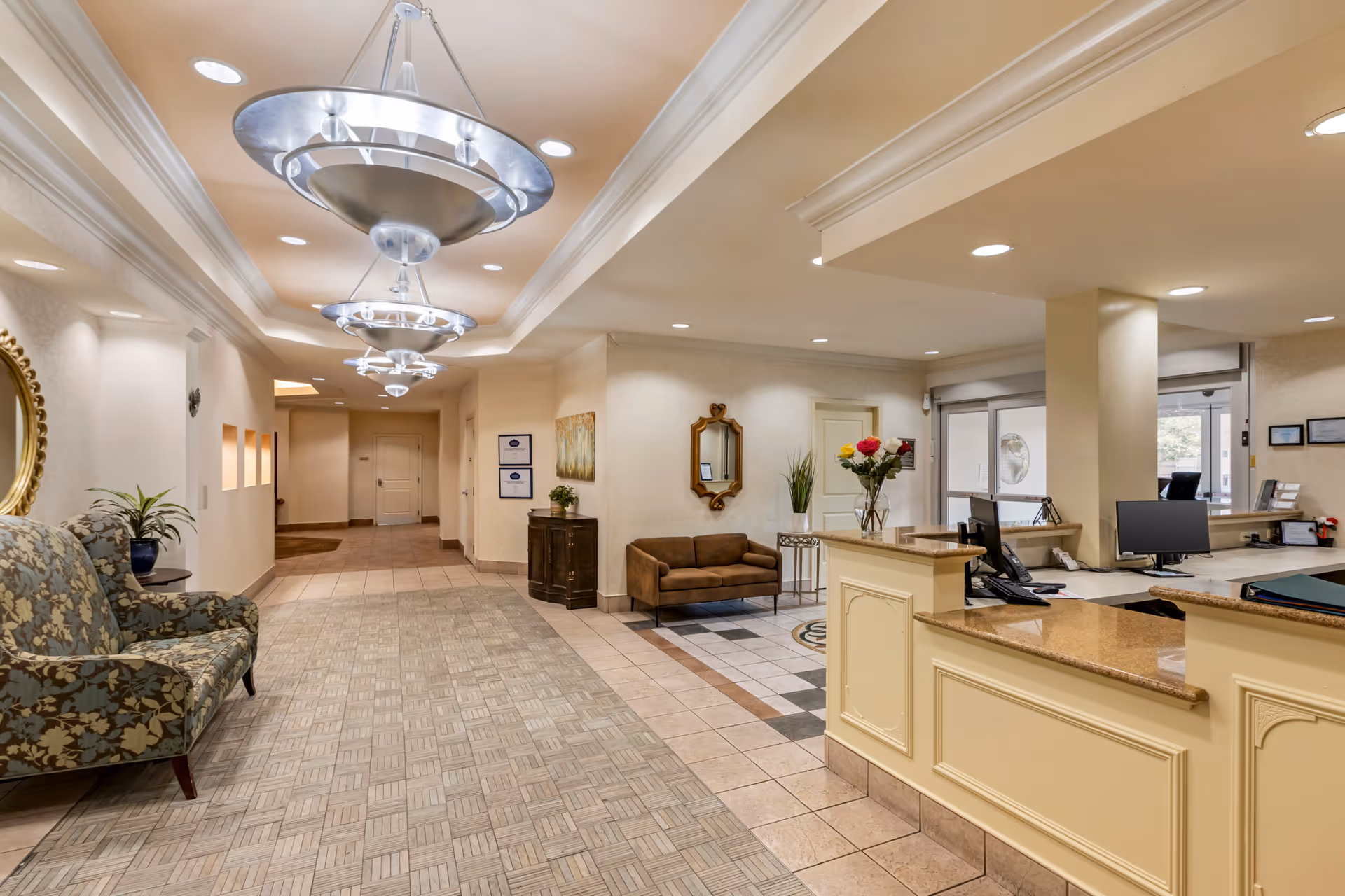 Reception area of Plum Creek Chicago featuring a front desk with computer monitors, a vase with flowers, a brown sofa, a patterned armchair, decorative mirrors on the walls, and modern ceiling lights.