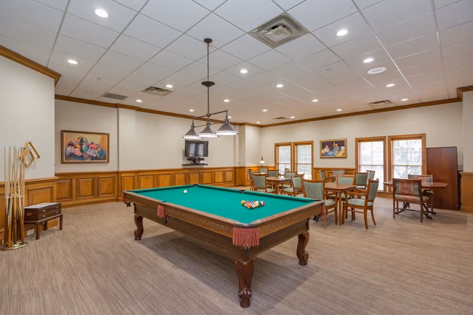 A spacious recreational room with a green felt pool table in the center, surrounded by wooden chairs and tables. The room has beige walls with wooden paneling, several framed paintings, and large windows letting in natural light. Ceiling lights and a hanging light fixture illuminate the space.