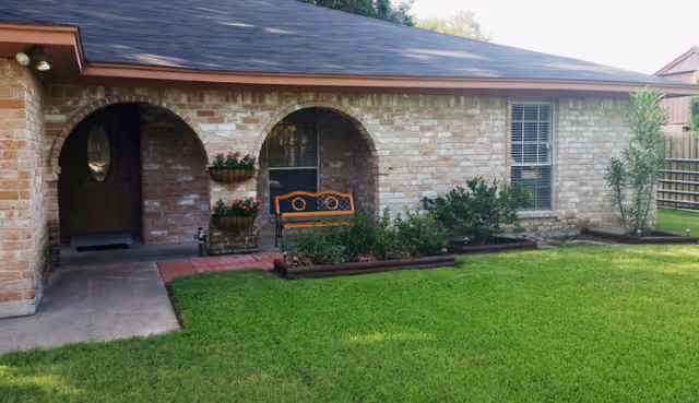 Exterior view of a single-story brick building with a brown roof. The entrance features two arched doorways, one with a door and the other with a bench and flower pots mounted on the wall. There is a well-maintained green lawn and some shrubs in front of the building.