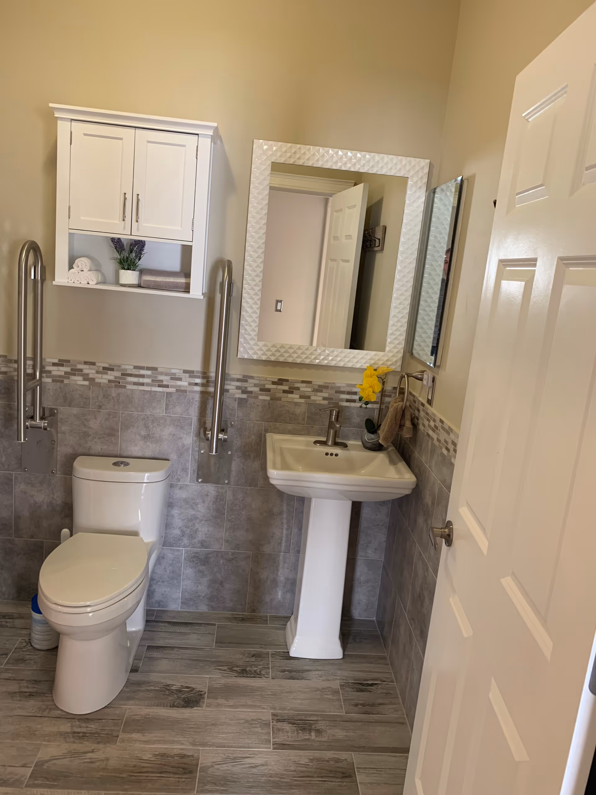 A clean and modern bathroom with a white toilet and a white pedestal sink. There are two stainless steel grab bars on either side of the toilet. Above the toilet is a white wall cabinet with two doors and an open shelf holding rolled white towels, a small potted plant, and a folded towel. A large rectangular mirror with a textured white frame hangs above the sink. The walls are tiled halfway up with gray and beige tiles, and the floor has wood-look gray tiles. A yellow flower in a small vase is placed on the sink.