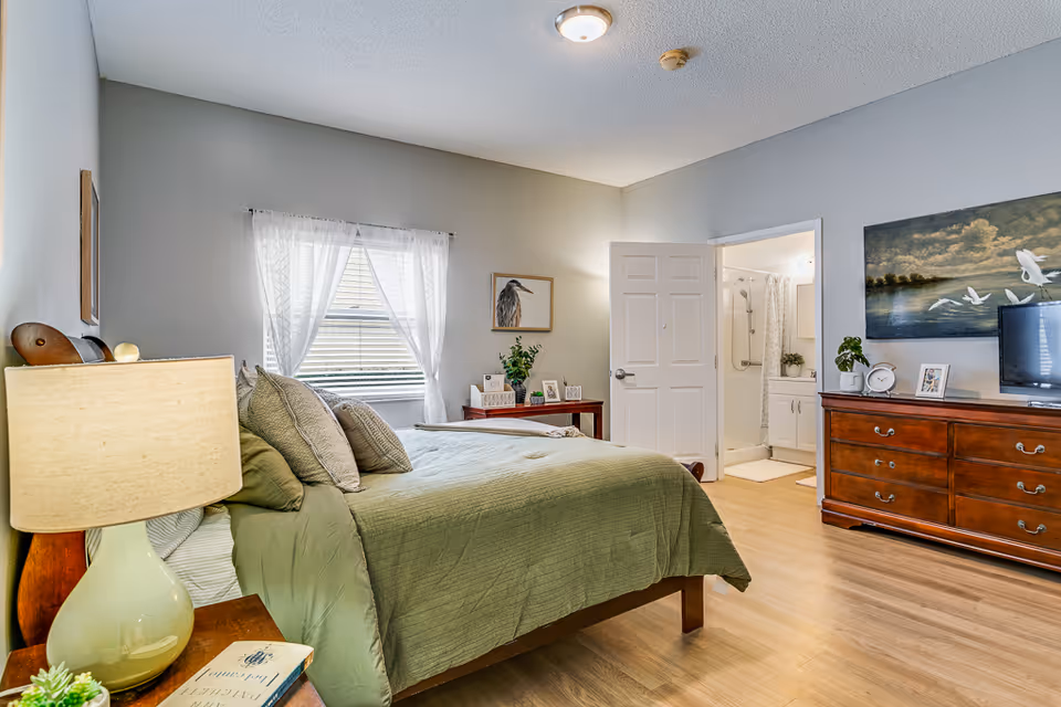 Well-lit bedroom with a green-covered bed, bedside lamp, dresser with a TV, and an open door to a bathroom.