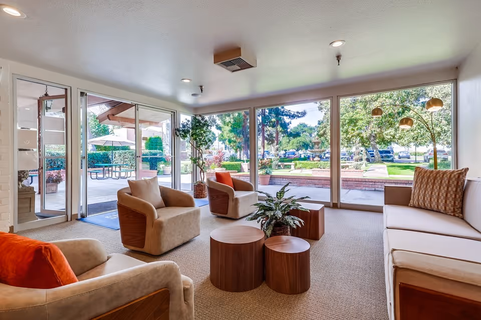 Bright senior living lounge with armchairs, a sofa, wooden coffee tables and large windows overlooking a landscaped courtyard.