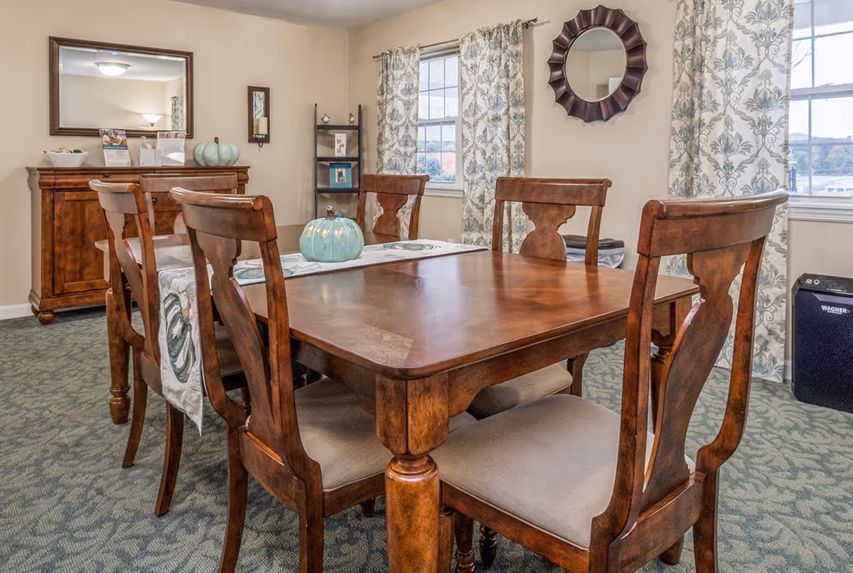 A dining room with a wooden dining table and six matching chairs. The table has a decorative runner and a ceramic pumpkin centerpiece. The room has patterned curtains on two windows, a wooden sideboard with a large mirror above it, and a small shelving unit with framed photos. The carpet is patterned, and a decorative round mirror hangs on the wall.