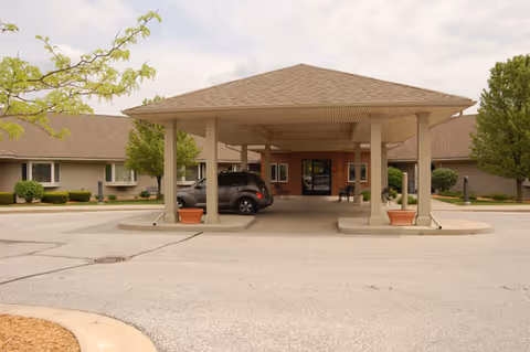 Entrance of a senior living facility with a covered drop-off area supported by columns. A black SUV is parked under the canopy. The building has a beige exterior with a brown roof and some greenery around the perimeter.