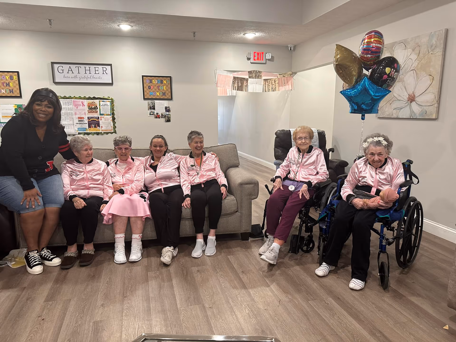 A group of elderly women and a younger woman sitting together in a living room area. Five elderly women are wearing matching pink satin jackets, with two of them seated in wheelchairs. The younger woman is sitting on the left side, wearing a black jacket and denim shorts. There are colorful balloons on the right side and a wall decoration with the word 'GATHER' above a bulletin board in the background.