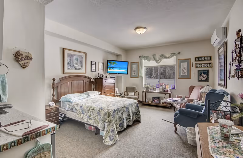 A cozy senior living bedroom with a wooden bed covered in a floral quilt, a wooden dresser, and a wall-mounted TV displaying a beach scene. The room has a window with sheer curtains, several framed pictures on the walls, and comfortable seating including a pink armchair and a blue recliner. There is a small table with family photos and a lamp, and a kitchenette area with a countertop and sink is partially visible on the left.