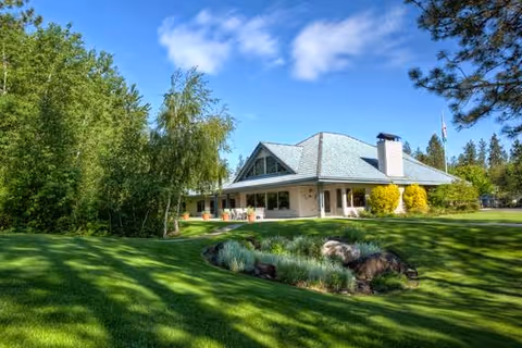 A single-story clubhouse-style building with a sloped roof on a well-manicured lawn surrounded by trees under a blue sky.