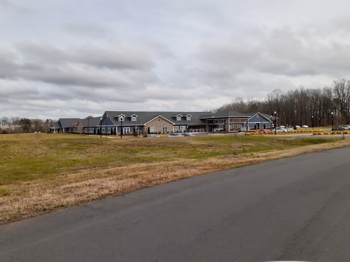 Wide exterior view of a single-story nursing and rehabilitation center building with a large grassy area and a paved road in the foreground under a cloudy sky.