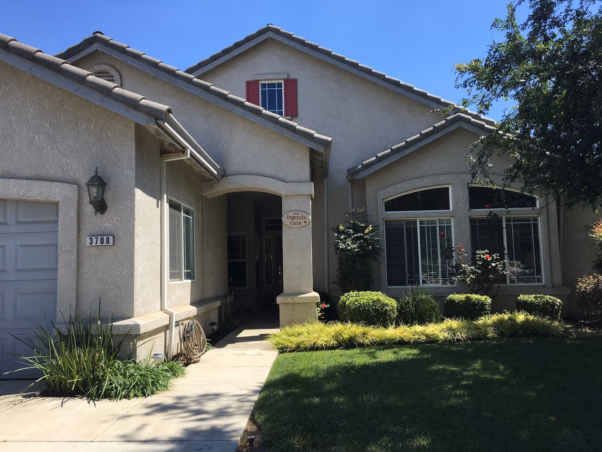 Exterior view of a single-story residential building with beige stucco walls and a tiled roof. The house has a garage on the left, a covered entrance in the center, and large windows with white blinds on the right. There is a well-maintained lawn and various shrubs and plants in front of the house. A tree is visible on the right side, and the sky is clear and blue.