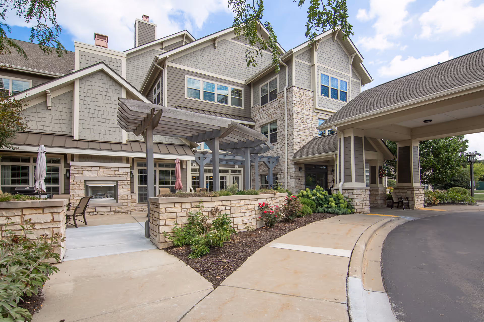 Exterior view of a senior living facility with stone and siding facade, a covered entrance driveway, outdoor seating area with umbrellas, and landscaped plants and flowers under a partly cloudy sky.