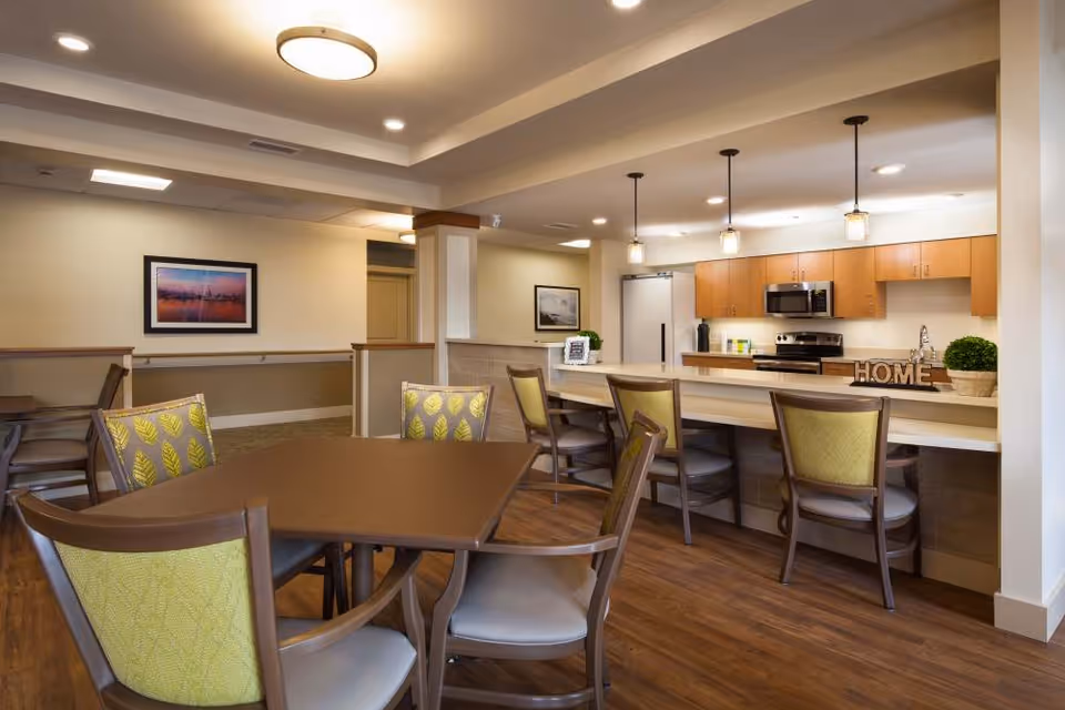 A bright and modern dining area with a wooden table and chairs featuring green patterned upholstery. In the background, there is a kitchen with wooden cabinets, stainless steel appliances, and a countertop with bar stools. The room is well-lit with ceiling lights and pendant lights above the kitchen counter. Decorative elements include framed pictures on the walls and a small plant on the counter next to a wooden 'HOME' sign.