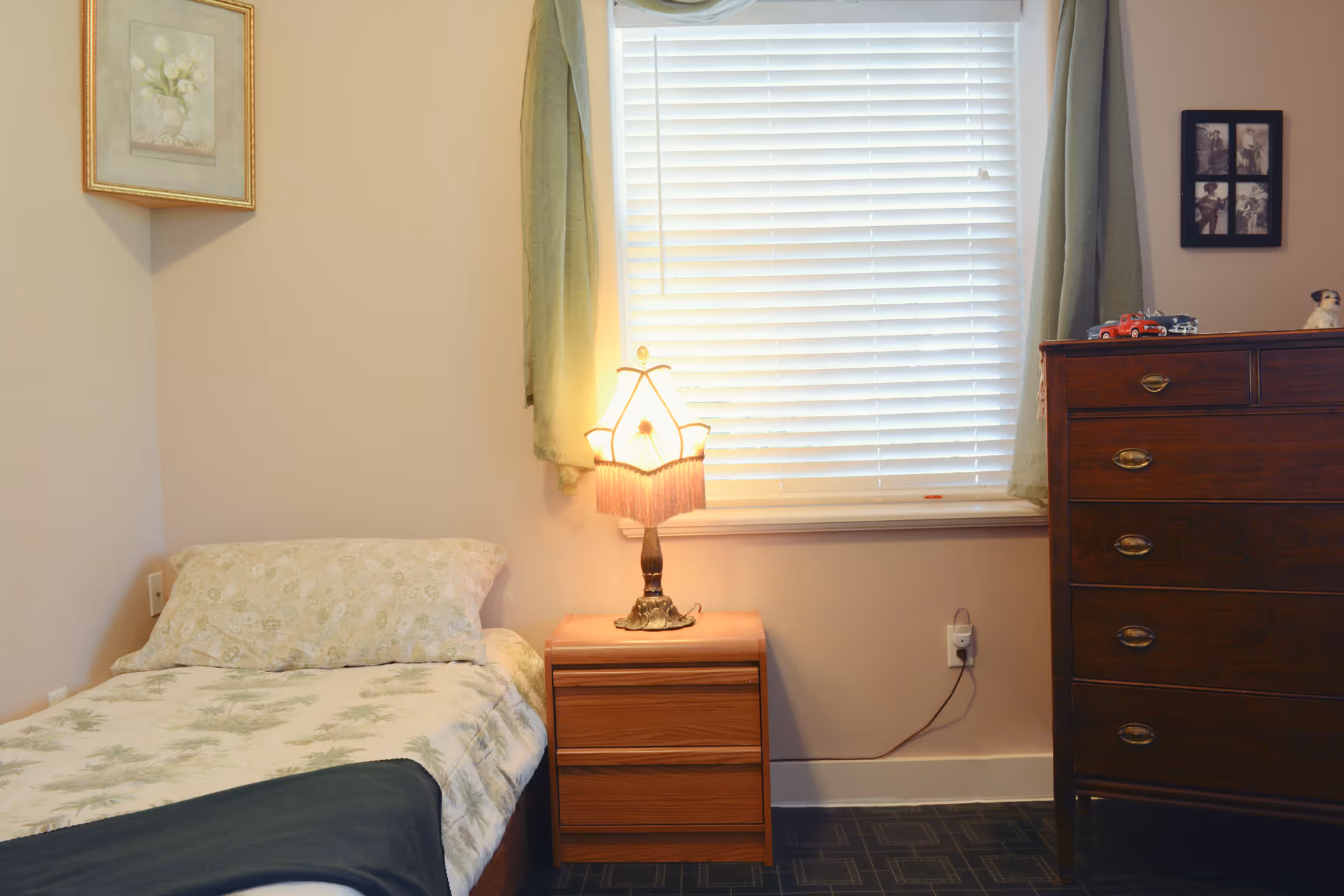 Small bedroom with a single bed, a wooden nightstand holding a lamp beneath a window with blinds, and a tall dresser.