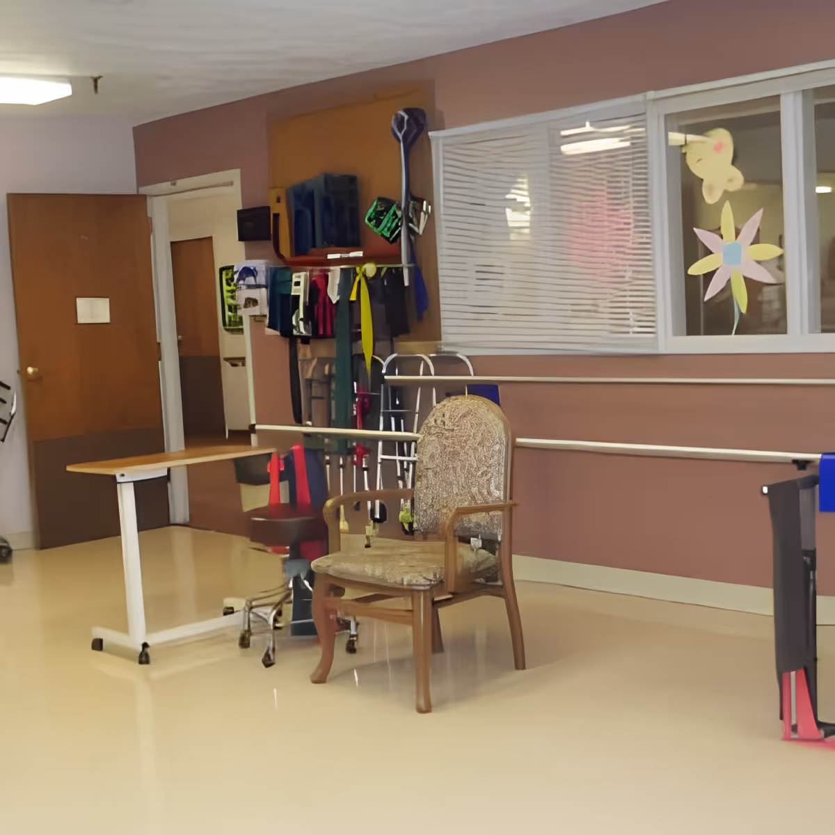 A senior facility common area with a padded armchair, walkers and mobility equipment, handrails along the wall, and a small overbed table.