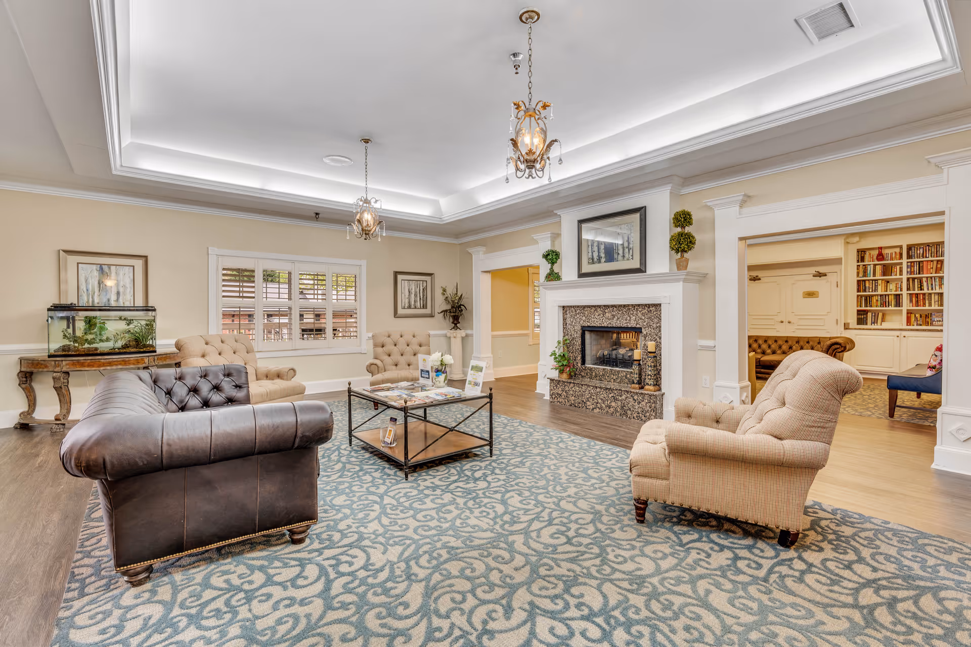 A spacious and elegant living room in a senior living facility featuring a large patterned area rug, a dark leather tufted sofa, three beige tufted armchairs, a glass coffee table with brochures, a fireplace with a granite surround, decorative plants, framed artwork, and built-in bookshelves visible through an open doorway.