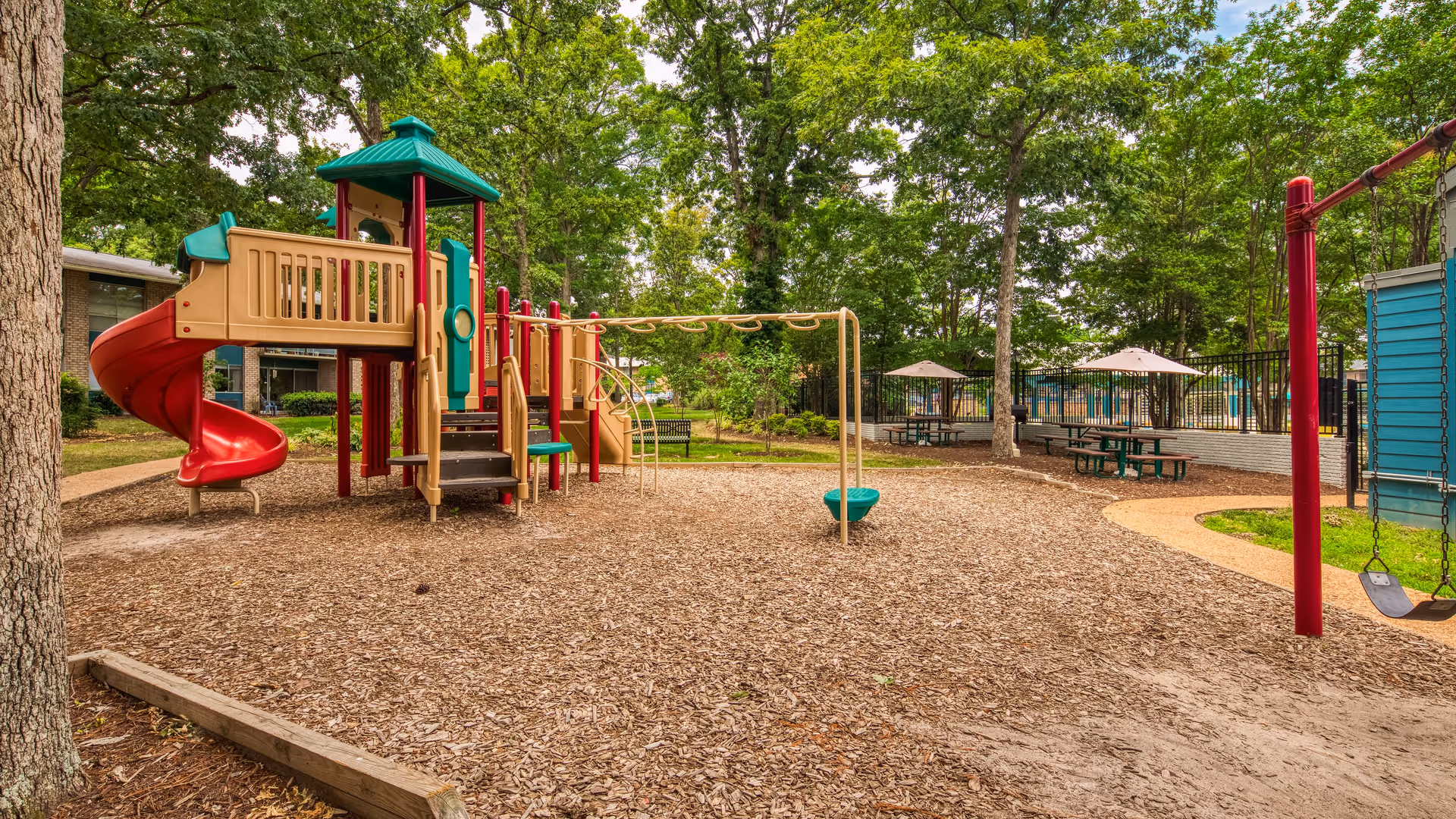 Outdoor playground area with a red spiral slide, climbing structure, monkey bars, and swings surrounded by wood chips. There are picnic tables with umbrellas and trees in the background, along with a building partially visible on the left side.