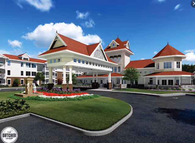 Exterior view of a senior living facility named Victoria In McKinney featuring a large covered entrance with red roofs, white walls, and landscaped gardens with flowers and greenery under a blue sky with clouds.