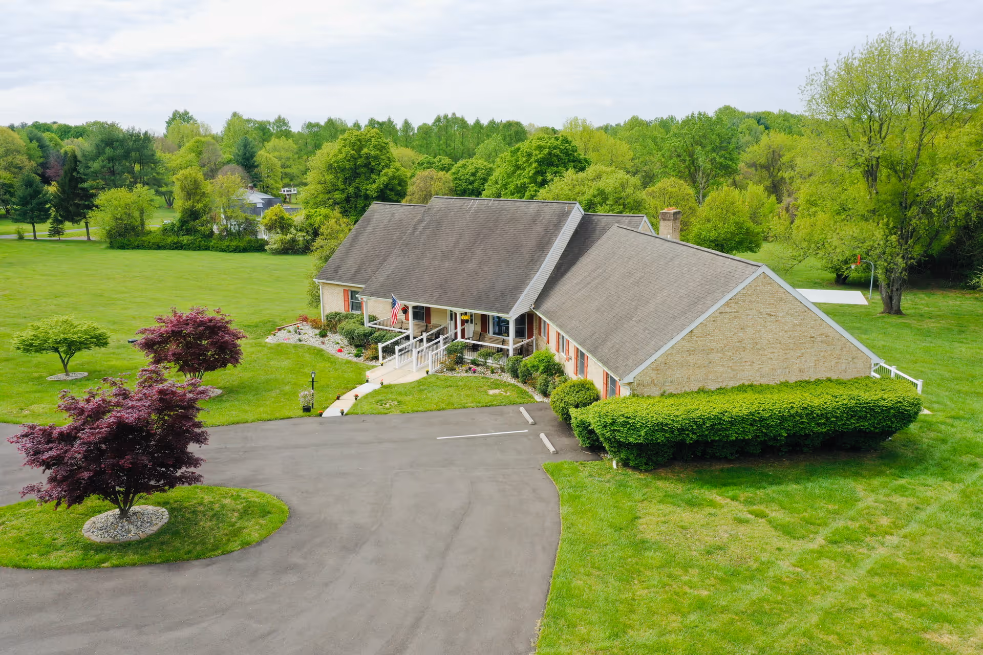 Aerial view of a single-story building with a sloped roof surrounded by green lawns and trees. The building has a ramp leading to the entrance, an American flag near the door, and a paved driveway with parking spaces. The area is lush with greenery and appears peaceful.