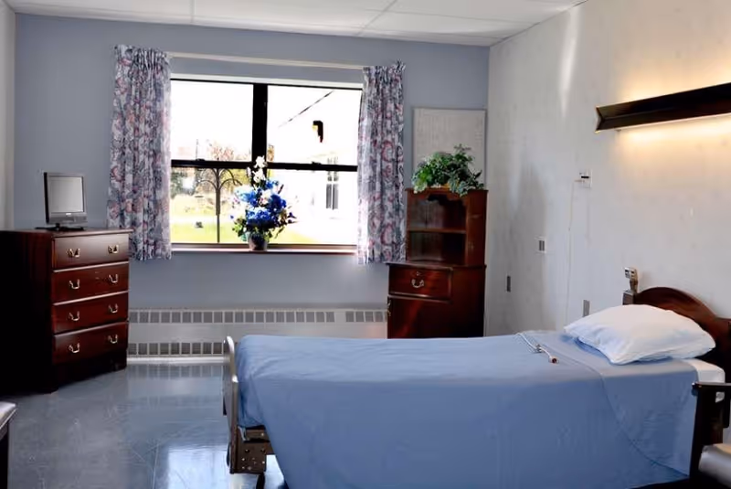 A simple, clean bedroom in a nursing facility with a single bed covered in a blue blanket, a pillow, and a call button on the bed. There is a window with floral curtains letting in natural light, a wooden dresser with a small TV on top, and a wooden cabinet with a plant on it. The walls are light-colored and there is a wall-mounted light fixture above the bed.