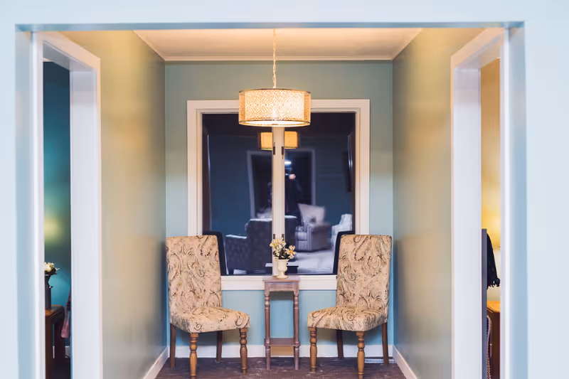 Two upholstered chairs flank a small side table beneath a hanging light in a cozy seating nook with a window.