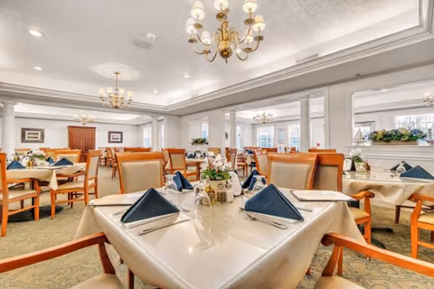 Spacious formal dining room with multiple tables set with folded navy napkins, floral centerpieces, chandeliers and upholstered chairs.