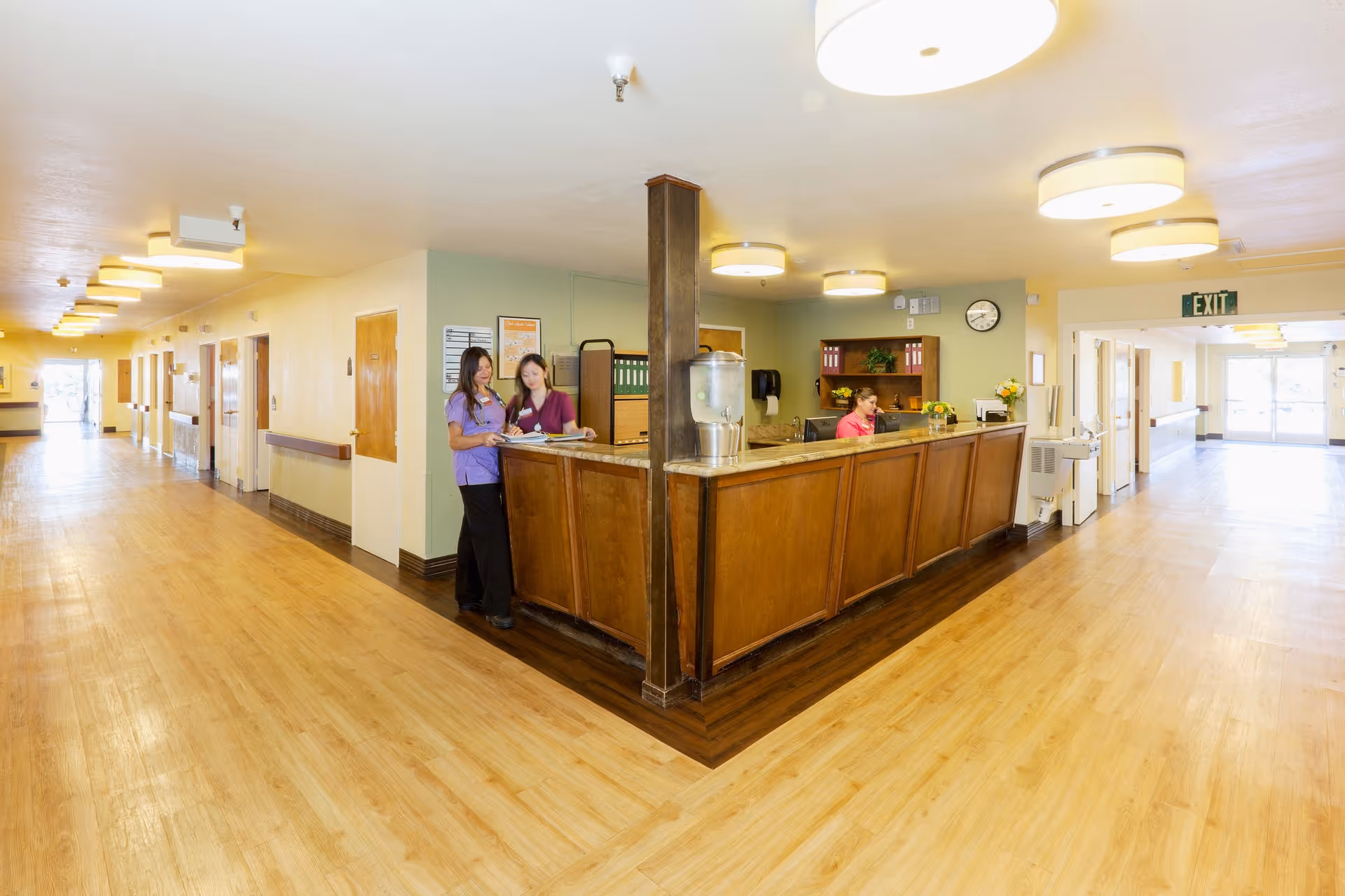A bright hallway in a healthcare facility with wooden floors and light-colored walls. In the center is a wooden reception desk with a water dispenser on it. Behind the desk, a woman is seated working on a computer. Two women in purple and maroon scrubs are standing and talking near the desk. The hallway extends in both directions with doors along the walls and ceiling lights evenly spaced.