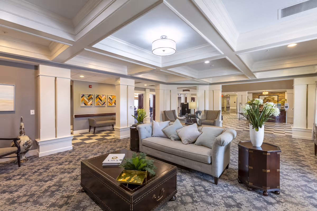 A spacious and well-lit living room area in a senior living facility with a light gray sofa adorned with multiple cushions, two armchairs, a dark wooden coffee table with decorative items, and a side table with a large white vase filled with white flowers. The room features a coffered ceiling with recessed lighting, patterned carpet flooring, and several columns. In the background, there is a hallway with additional seating and artwork on the walls.