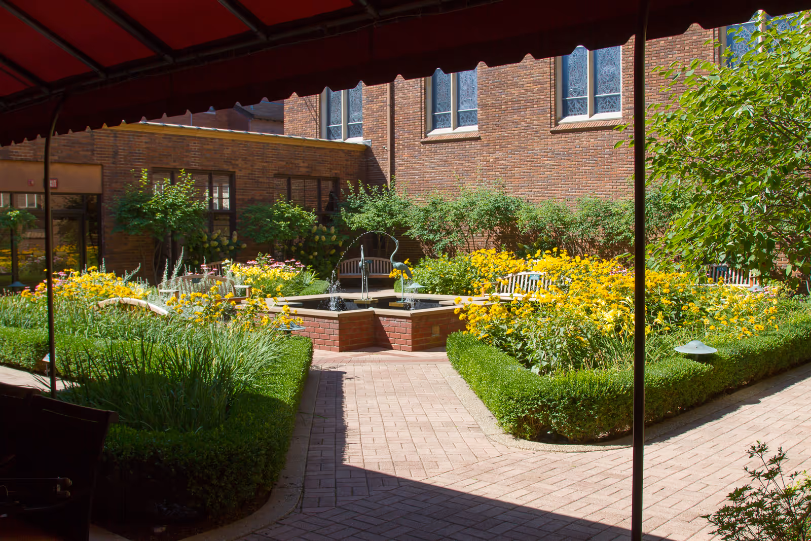 A peaceful outdoor garden area with a brick-paved walkway, neatly trimmed hedges, vibrant yellow flowers, and a small central fountain surrounded by benches. The garden is enclosed by brick building walls with windows.