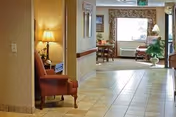 A hallway in a senior living facility with tiled floors and beige walls. On the left side, there is a wooden chair with a lamp on a small table behind it. At the end of the hallway, there is a seating area with a window letting in natural light, a table with chairs, a lamp, and some plants.