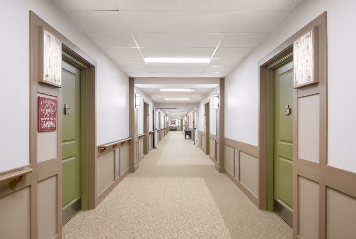A long, well-lit hallway in a senior living facility with beige walls and carpet. The hallway has green doors on both sides, each with a small plaque. There are handrails along the walls and decorative light fixtures above the doors. A sign on the left side reads, 'If you don't believe in angels you have not met my grandma.'