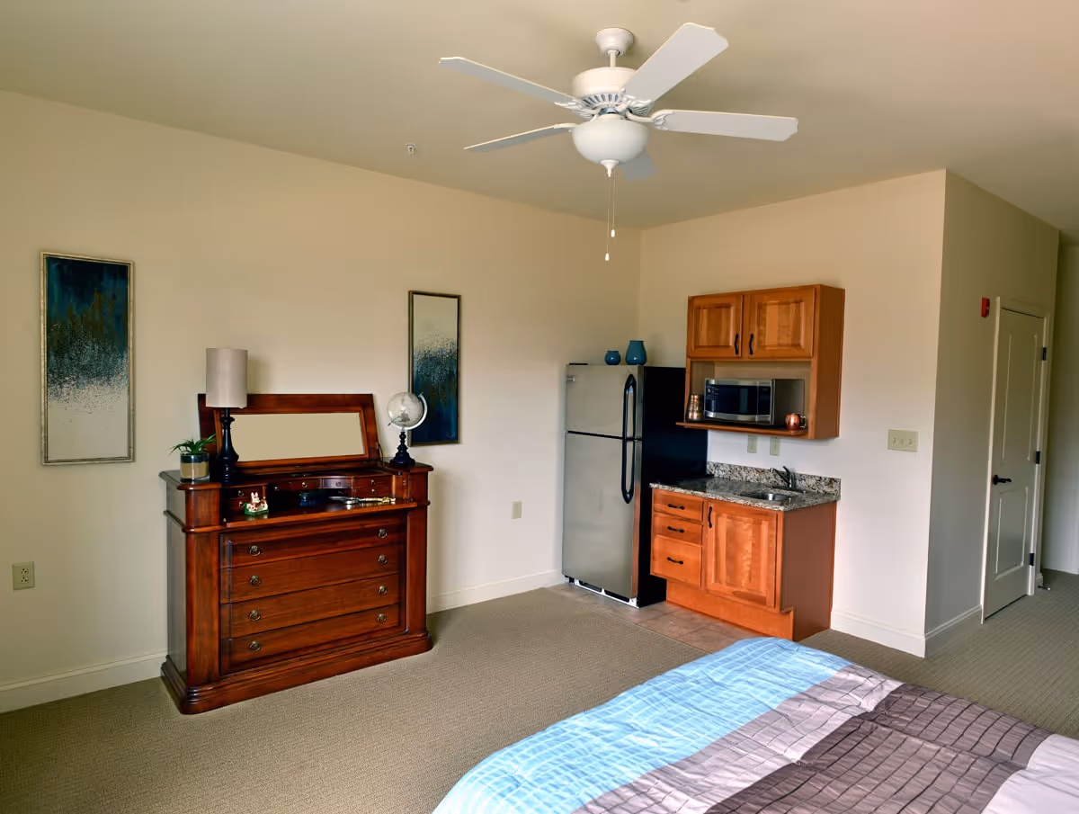 Interior view of a senior living facility room featuring a bed with a blue and brown striped comforter in the foreground, a wooden dresser with a mirror, lamp, and decorative items on top, two abstract paintings on the wall, a ceiling fan, and a kitchenette area with a small refrigerator, microwave, sink, and wooden cabinets.