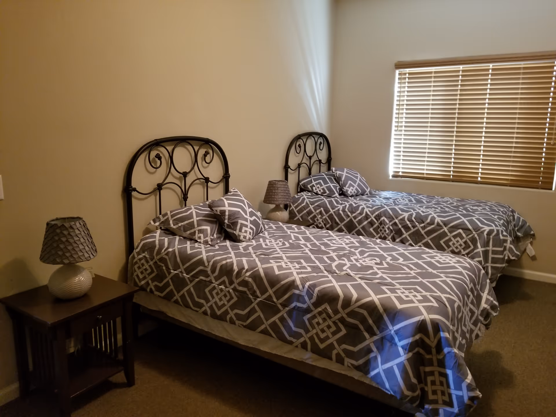 A bedroom with two twin beds featuring black metal headboards and matching gray and white geometric patterned bedding. Each bed has two pillows. There is a wooden nightstand between the beds with a decorative lamp on it. A window with closed blinds is on the right wall.