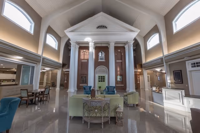 Spacious and elegant interior of a senior living facility with high vaulted ceilings, large white columns, and a brick wall backdrop. The room features comfortable seating including sofas and armchairs arranged around a small table, with additional tables and chairs along the sides. Large arched windows allow natural light to fill the space.