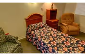 Small single-occupancy bedroom with a floral bedspread, wooden headboard, nightstand, lamp, and two chairs.