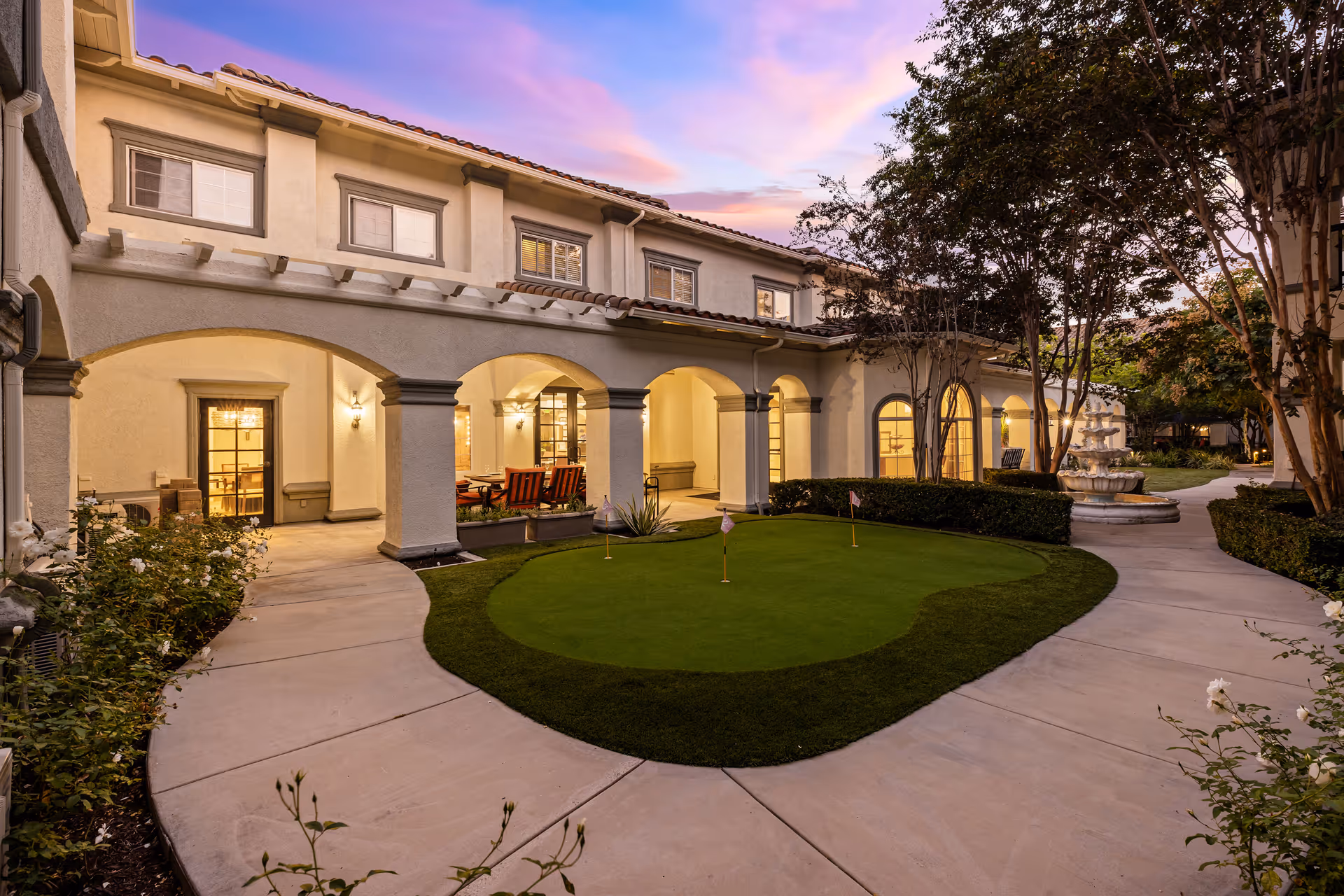 Dusk courtyard with arched colonnade, small putting green, and a fountain outside a two-story building.