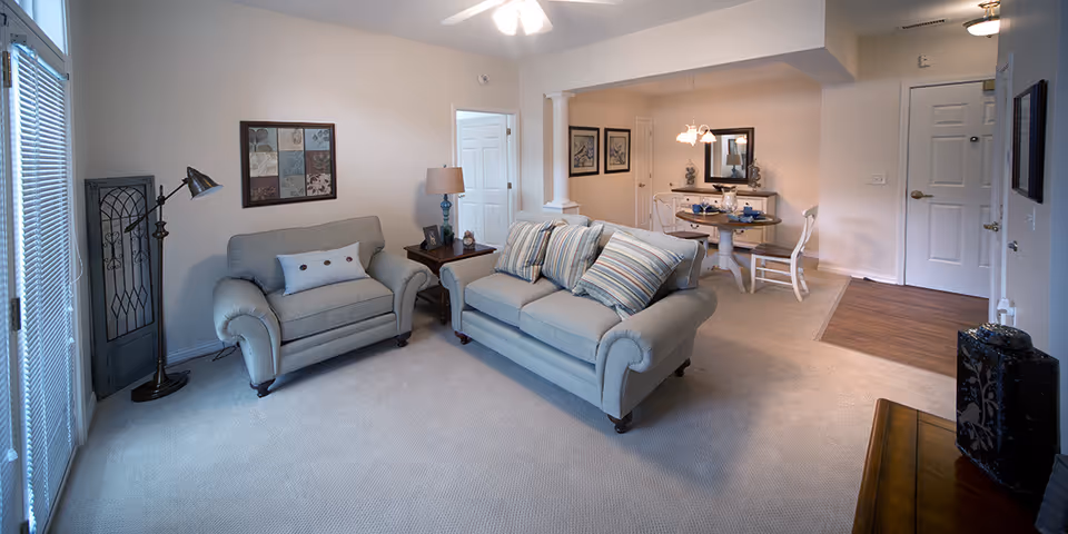 A cozy living room area with a light gray loveseat and matching armchair, both adorned with decorative pillows. A floor lamp stands next to the armchair, and a framed artwork hangs on the wall above it. In the background, there is a small dining area with a round table set for two, a mirror, and two framed pictures on the wall. The room has beige carpeting and light-colored walls, with a door and a window with blinds visible.