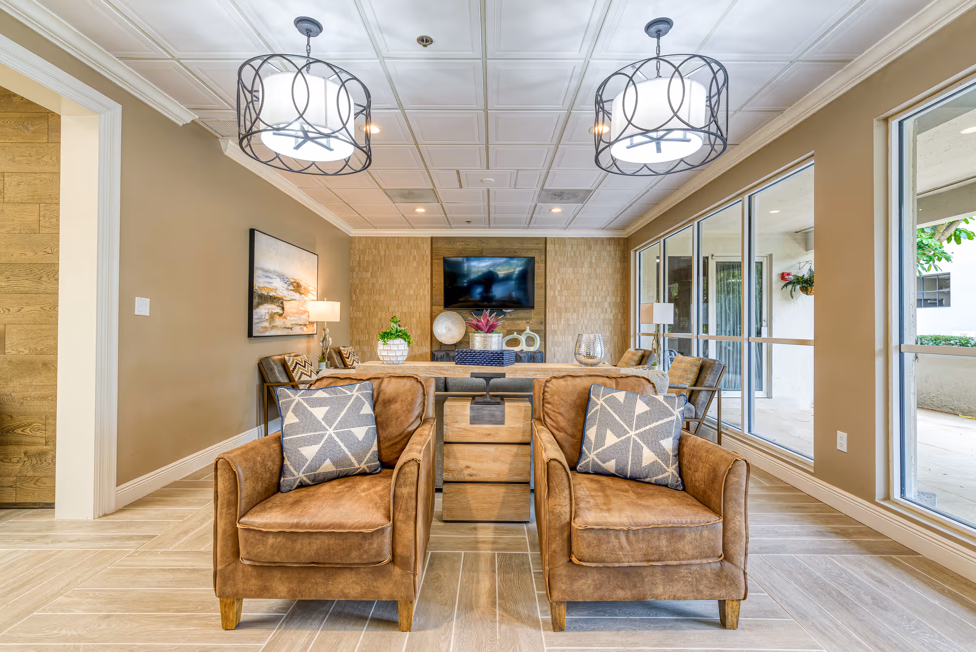 A cozy senior living room with two brown leather armchairs featuring geometric patterned cushions in the foreground. Behind the chairs is a wooden side table and a sofa with decorative plants and ornaments on it. The room has large windows on the right side letting in natural light, two modern pendant lights hanging from a white tiled ceiling, and a flat-screen TV mounted on a textured wall at the back.