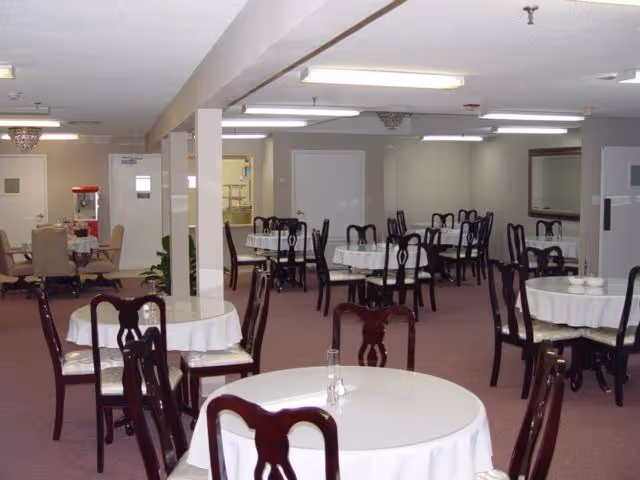 A dining room with multiple round tables covered with white tablecloths and dark wooden chairs arranged around them. The room has a carpeted floor, fluorescent ceiling lights, and a popcorn ceiling. There is a popcorn machine visible in the background near a doorway.