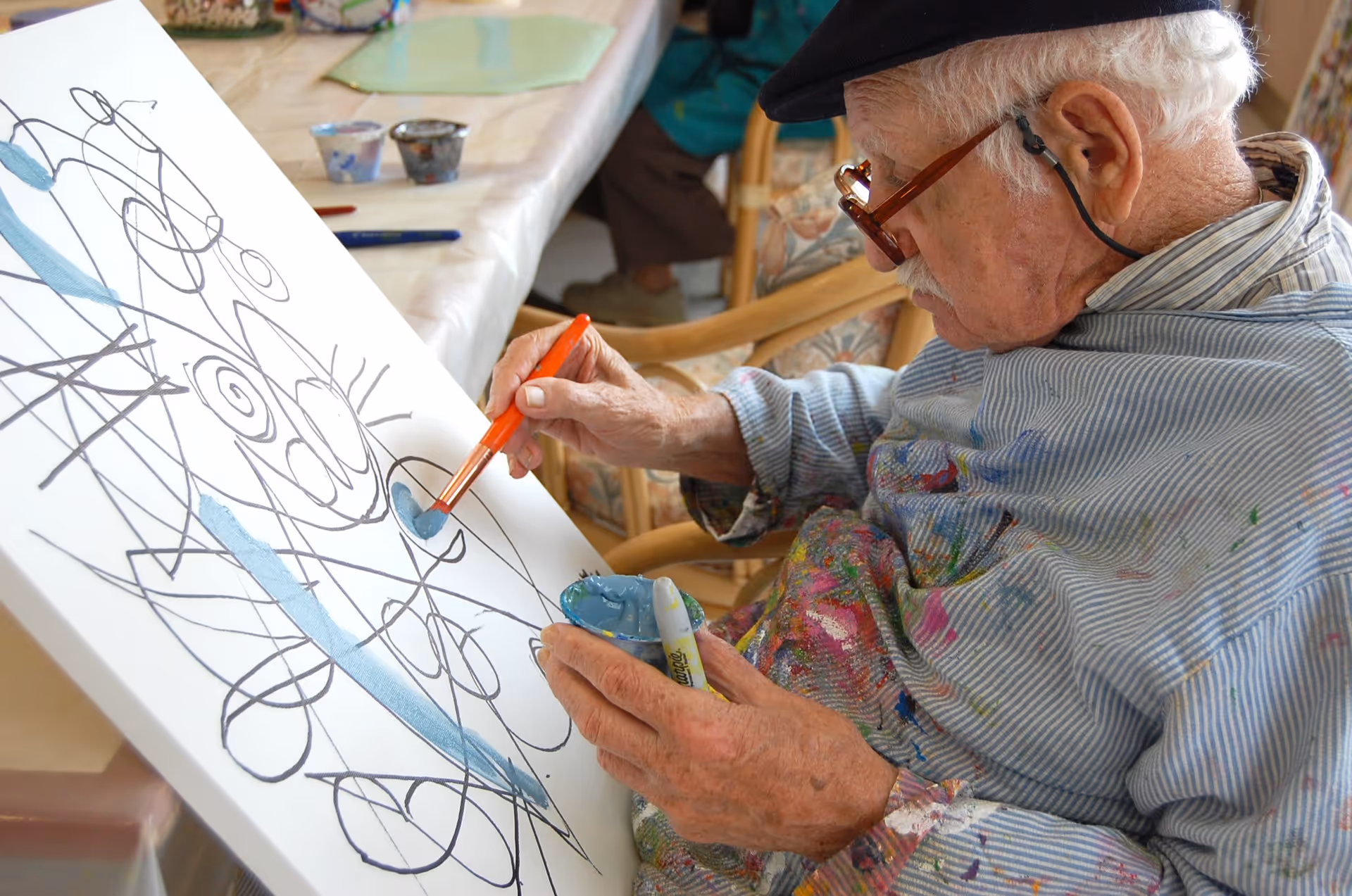 An elderly man wearing glasses and a paint-splattered smock is painting on a canvas with abstract black lines, using a paintbrush dipped in blue paint. He is seated at a table with art supplies in the background.