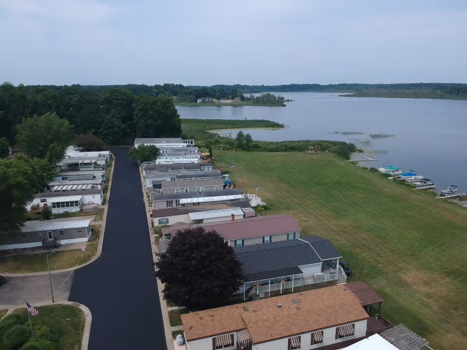 Aerial view of a waterfront community with multiple single-story homes lined along a paved road. The community is adjacent to a large lake with several boats docked along the shoreline. There is a large grassy area between the homes and the water, and dense trees are visible in the background.