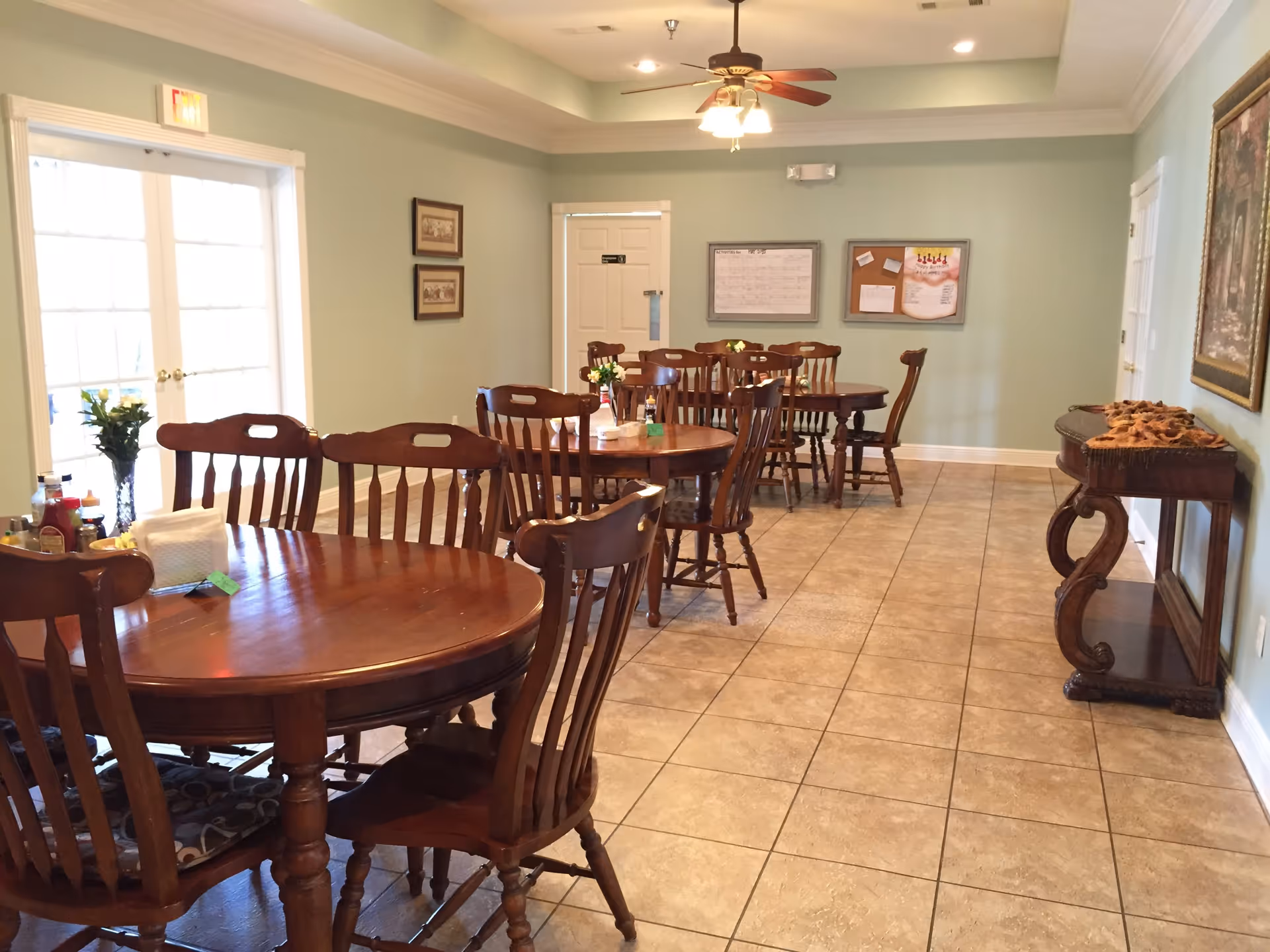 A bright dining room with several round wooden tables and chairs on a tiled floor under a ceiling fan.