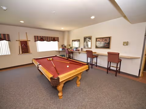 A recreational room with a pool table in the center, featuring a red felt surface and pool cues resting on it. The room has beige walls, carpeted floor, and recessed ceiling lights. On one side, there is a long counter with three high chairs and framed pictures hanging above it. A window with a valance is visible on the adjacent wall, along with a wall-mounted rack holding additional pool cues.