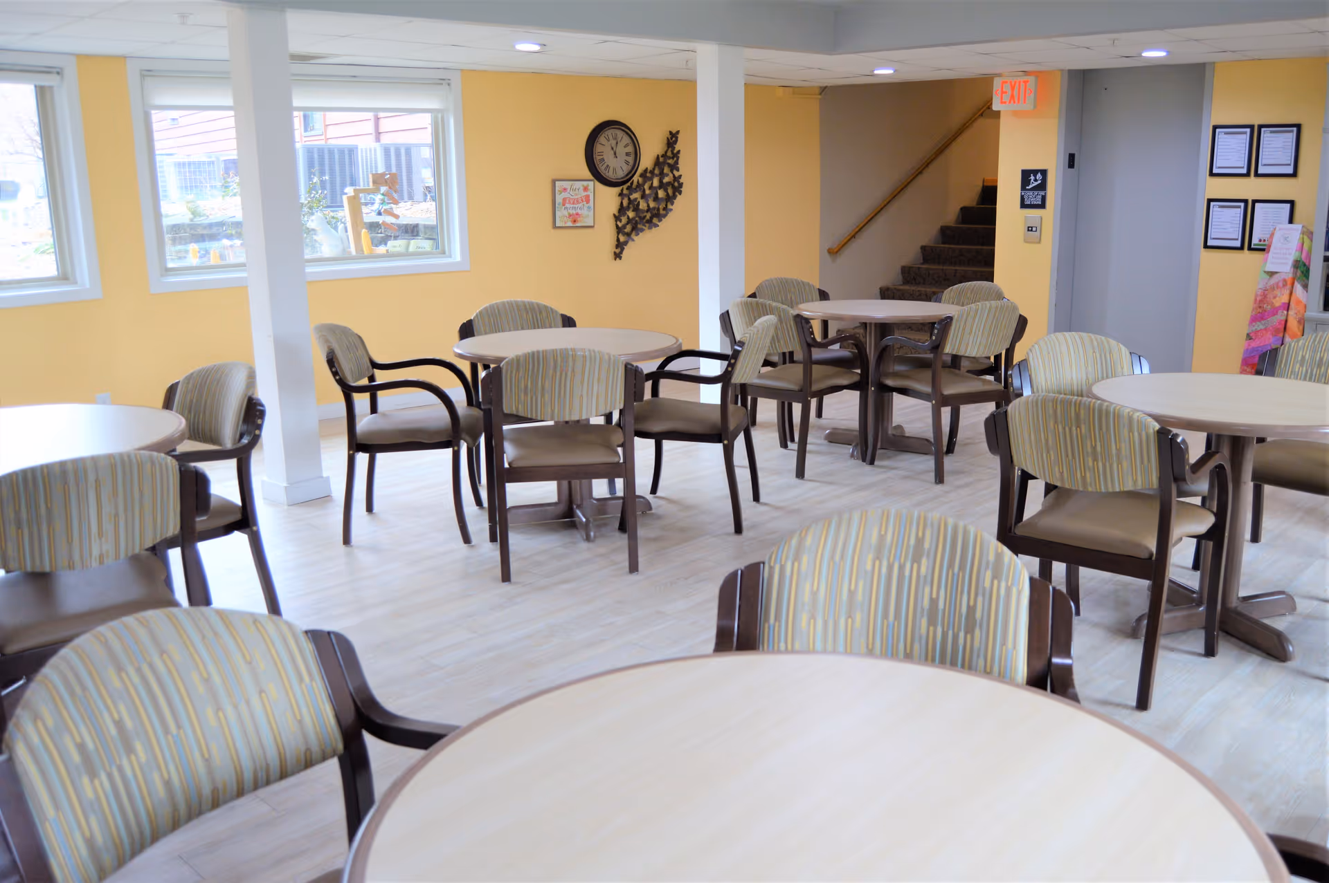 A bright room with multiple round tables and cushioned chairs arranged around them, yellow walls, two windows letting in natural light, a clock and wall decorations, and a staircase leading up in the background.