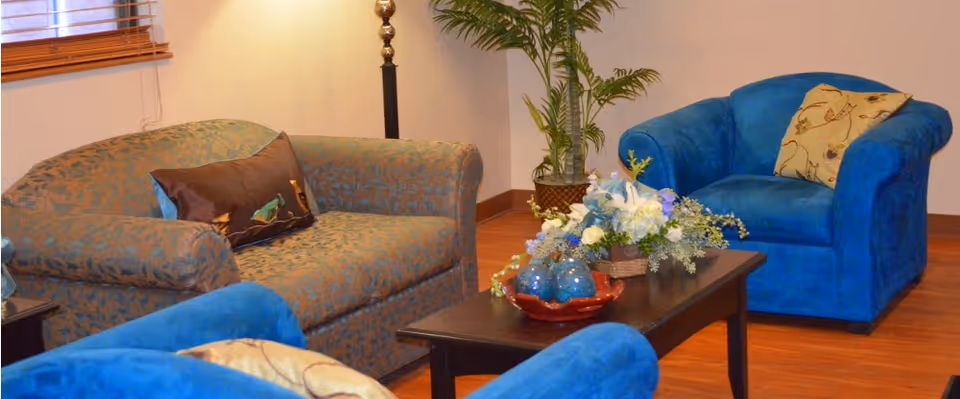 A cozy living room area with a patterned sofa, two blue armchairs, a wooden coffee table with a floral arrangement and decorative items, a tall lamp, a potted plant, and a window with wooden blinds.