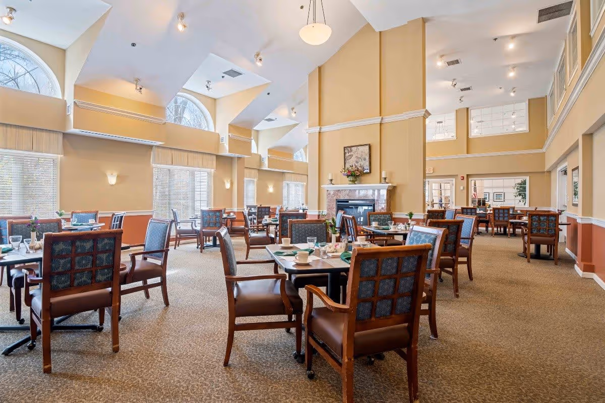 A spacious dining room with multiple tables and chairs arranged neatly. The room features high ceilings with large arched windows allowing natural light to fill the space. There is a fireplace with a floral arrangement and artwork above it. The tables are set with cups, glasses, and napkins, ready for a meal.